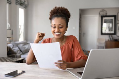 Neşeli genç Black serbest çalışan profesyonel bir kadın, kazanan jest yapıyor, evet el iş yerinde dizüstü bilgisayarla oturuyor, finansal kağıt dökümanları okuyor, büyük haberlerle fark ediyor, gülümsüyor, gülüyor.