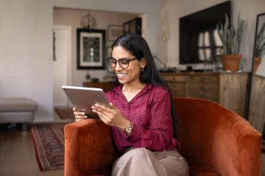 Mutlu genç Hintli kadın tabletten içeriği izliyor veya okuyor, evinde koltuğunda oturuyor, alet kullanıyor, gülümsüyor, gülüyor, internetten keyif alıyor, kablosuz iletişimden keyif alıyor.