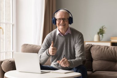 Emekliliğimde kazanmayı öneriyorum. Neşeli, mutlu, yaşlı, yaşlı, kulaklıklı uzaktan kumandalı bir öğrenci dizüstü bilgisayardan dikkati dağılmış bir şekilde kameraya baş parmağını gösteriyor.