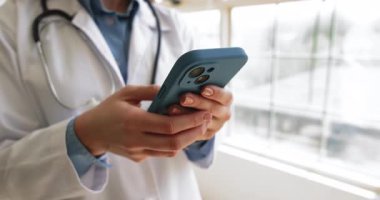Paltolu kadın doktor tatilde pencerenin yanında duran akıllı telefonu kullanıyor, kişisel veya hasta mesajlarına cevap veriyor, elektronik sağlık kayıtlarına erişiyor, internette tıbbi bilgilere erişiyor.
