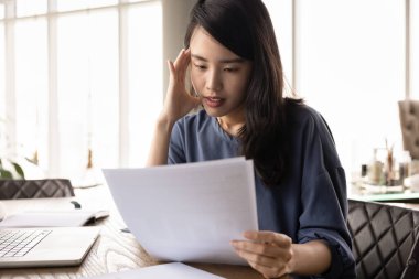 Ciddi, güzel, Asyalı genç bir kadın ofis işyerinde kağıt okuyor, kafaya dokunuyor, sorunlar yaşıyor, maddi sorunlar yaşıyor, para kaybı, haberlerle ilgili öğrenim görüyor.