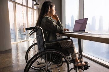 Ciddi bir iş projesi yöneticisi, felçli bir kadın tekerlekli sandalyede dizüstü bilgisayarda oturuyor, online içeriği analiz ediyor, çağdaş ofiste tek başına büyük pencerelerle çalışıyor.