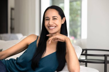 Neşeli, genç ve güzel Yeni Zelandalı kadın portre için rahat bir evde poz veriyor, yumuşak koltukta dinleniyor, kameraya dişlek tebessümle bakıyor, çeneye dokunuyor, keyfine bakıyor, rahatlıyor, dinleniyor, mola veriyor.