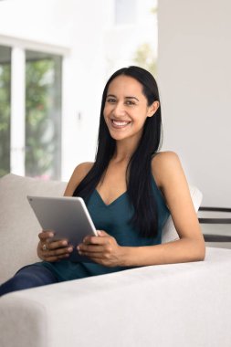 Neşeli genç Maori kadın dijital tablet üzerinde yumuşak ev kanepesinde online servis kullanıyor, dişlek tebessümle kameraya bakıyor, internet iletişiminden hoşlanıyor, dikey portre için poz veriyor.