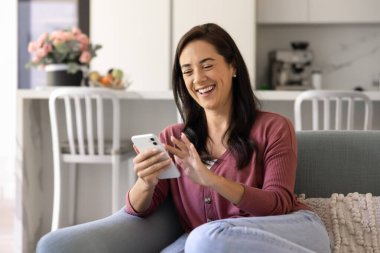 Kanepede oturmuş gülümseyen ve rahat oturma odasında akıllı telefon kullanan, iyi haberlerle, mesajlarla, mesajlarla, sosyal medyada gezinen bir kadın. Dijital iletişim, teknoloji ve boş zaman