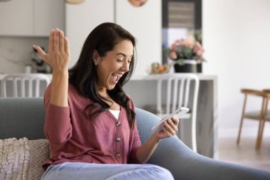 Kanepede oturan neşeli kadın, akıllı telefona şaşkın bir ifadeyle bak ve elini kaldır, çevrimiçi piyango zaferini, kişisel başarı haberlerini, teklifi, olumlu mesajı ya da başarıyı kutla.