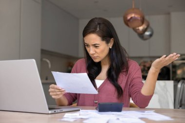 Endişeli orta yaşlı kadın elinde kağıt makbuzu, elinde dizüstü bilgisayar ve hesap makinesiyle masada oturuyor. Faturalar, vergiler ya da finansal sorunlar yüzünden hayal kırıklığına uğramış. Borç konusunda stresli görünüyor.