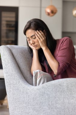 Evde koltukta oturan yorgun bir kadın, gözleri kapalı bir şekilde başını tutuyor, stres, depresyon, şiddetli baş ağrısı, zihinsel sağlık sorunları, duygusal mücadele. Migren, kan basıncı.
