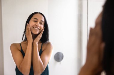 Mutlu, genç, güzel yerli Maori kadın aynaya kapalı gözlerle dokunuyor, gülümsüyor, güzellik bakımının keyfini çıkarıyor, cilt bakımının keyfini çıkarıyor, pürüzsüz, temiz yüz derisini okşuyor.