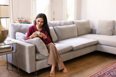 Pozitif genç Hintli ev sahibi kadın cep telefonuyla sosyal medyaya kayıyor, akıllı telefonuyla daktilo çekiyor, rahat rahat ferah koltukta oturuyor şık oturma odasının iç dekorasyonunda.