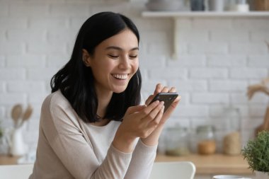 Fotoğrafta gülümseyen Asyalı kadın mutfakta akıllı telefon kullanıyor, mutlu genç bayan telefon ekranına bakıyor, sosyal ağda mesaj yazıyor, sohbet ediyor, cep telefonuyla evde vakit geçiriyor.