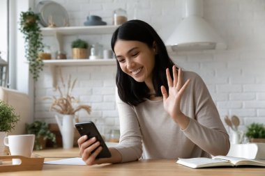 Gülümseyen Asyalı kadın, telefon kamerasına el sallıyor, mutfakta video görüşmesi yapıyor, akıllı telefon ekranına bakan mutlu genç bayan, internette sohbet ediyor, sanal bir toplantıda, konferansta.