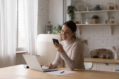 Mutlu, neşeli, serbest çalışan, internetten sohbet eden, iş yerindeki akıllı telefondan internet uygulamasını kullanan, telefon görüşmeleri yapan, ekrana gülümseyen, gülen, mutfakta dizüstü bilgisayarı olan kız.