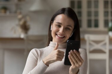 Video görüşmesi için kablosuz kulaklık ve cep telefonu kullanan mutlu genç bir kadın, ekranda el ele gösteriyor, konuşuyor, gülüyor, gülümsüyor, evde dinleniyor, internette konuşuyor.