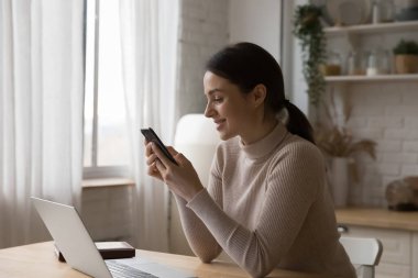 Mutlu odaklı cep telefonu kullanıcısı iş yerinde online sohbet ediyor, akıllı telefonda internet uygulaması kullanıyor, mutfak masasında dizüstü bilgisayarın başında oturuyor, iyi haberlerin tadını çıkarıyor.