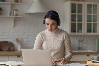 Kulaklıklı, internetten konferans dinleyen, proje ödevi yazan, rapor için notlar hazırlayan, evde sınava hazırlanan, mutfakta dizüstü bilgisayar başında çalışan ciddi derecede güzel bir kız öğrenci.