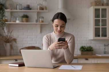 Bin yıllık serbest çalışan bir kız mutfakta akıllı telefon kullanıyor, internette sohbet ediyor, mesaj okuyor, laptopla çalışma masasında oturuyor. İletişim konsepti