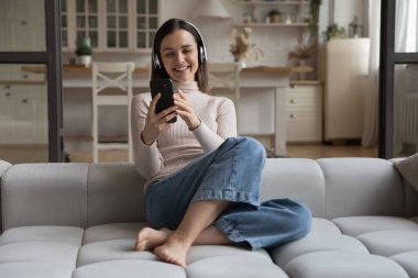 Kablosuz kulaklıklı neşeli kız akıllı telefondan konuşuyor, müzik dinliyor, şarkılar, melodiler dinliyor, internette medya içeriğini izliyor, gülüyor, evdeki kanepede gülümsüyor.