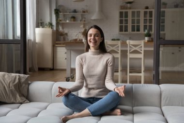 Mutlu, genç yogi kadın kanepede meditasyon yapıyor, lotus pozisyonunda oturuyor, elleri, parmakları zen tutuyor, gülüyor, gülümsüyor, kameraya bakıyor. Yoga, farkındalık konsepti. İçerideki portre