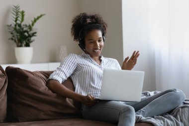 Kulaklıklı mutlu Afrikalı öğrenci evden video görüşmesi yapıyor, laptop kullanıyor, Webinar izliyor, çevrimiçi konferansa katılıyor, sanal sınıfa katılıyor, el sallıyor, web kamerasında konuşuyor, kanepede dinleniyor.