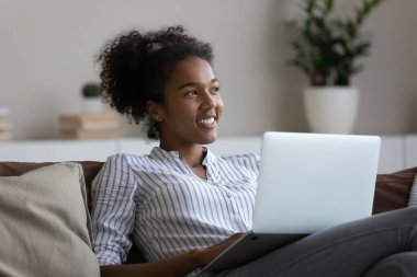 Düşünceli Afrikalı öğrenci kanepede dinleniyor, dizüstü bilgisayar kullanıyor, mutlu bir gülümsemeyle pencereye bakıyor, webinar öğrenmeyi, araştırma yapmayı, gelecekteki eğitimi, sanal eğitimi düşünüyor.