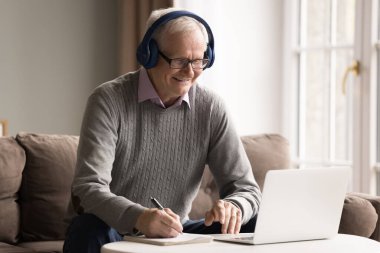 Aktif zihin. Modern kulaklıklı yaşlı, güler yüzlü bir erkek emekli ofis koltuğunda oturur, video kurslarını izler, not defterindeki çevrimiçi eğitimleri seyreder ve kağıt kopya defteriyle notlar alır.