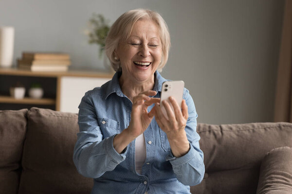 Digital laughter moment. Overjoyed stylish senior grandmother sit on sofa look at phone screen with candid laughter read funny message text make videocall shoot cute selfie picture scroll memes online