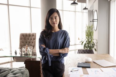 Kendine güvenen genç Asyalı pazarlama analisti ellerini çapraz yapıp kameraya bakıyor, ofis masasında portre için kağıt satış raporlarıyla poz veriyor.