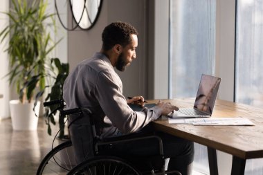 Tekerlekli sandalye kullanan, ofiste çalışan, iş yerinde dizüstü bilgisayarda yazan, pazarlama araştırma sonuçlarını analiz eden, satış raporları, online teknoloji kullanan ciddi Afrikalı Amerikalı iş adamı.