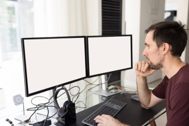 İki büyük ekranda yazı yazan, beyaz, yavan fotokopi alanı olan, çeneye dokunan, görevi düşünen serbest çalışan meşgul bir adam. Yazılım mühendisi, programcı, tüccar.