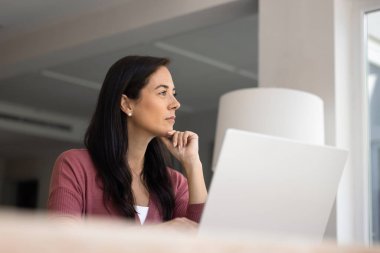 Ciddi bir orta yaşlı Latin kadın, gündelik giysiler içinde masada dizüstü bilgisayarla oturup düşünceli ve düşünceli bir ifadeyle mesafeye bakıyor. Yansıma, planlama, karar verme, uzak çalışma yaşam tarzı