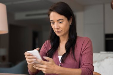 Çekici Latin kadın evde modern akıllı telefon kullanıyor, mesajları kontrol ediyor, mesajlaşıyor, online alışveriş yapıyor, sosyal medya paylaşımlarına göz atıyor, önemli haberleri okuyor veya odaklanmış ifadeyle bildiriyor