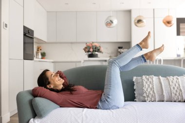 Neşeli bir kadın, ayakları havada, rahat bir kanepede uzanıyor, gülümsüyor, arka planda mutfağı olan çağdaş, modaya uygun bir oturma odasında tasasız anın tadını çıkarıyor. Akıllı bir evde boş ve rahat bir yaşam tarzı