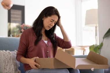 Kanepede oturan üzgün kadın karton paketi açıyor, yanlış paket kutusu alıyor, eksik sipariş yüzünden hayal kırıklığı yaşıyor, eksik eşyalar, orijinal ürün yerine düşük kalite veya sahte ürün. İade