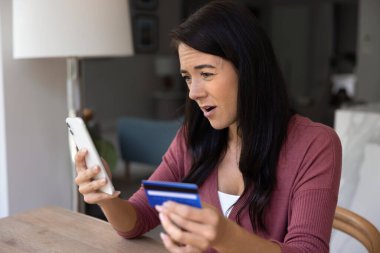 Kadın, kredi kartı ve akıllı telefonu tutarken endişeli görünüyor, çevrimiçi dolandırıcılık, dolandırıcılık, beklenmedik ödemeler, ödeme hatası, e-bankacılık sorunu veya kimlik doğrulama sorunları yüzünden asık suratlı.