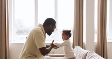 Aktif, iki ırklı bir kız, sevecen siyah baba ellerini tutuyor ve kanepeye zıplıyor, mutlu aile dansı yapıyor, evde birlikte eğleniyor. Çocuklarla vakit geçir, bağ kur ve babalık yap.