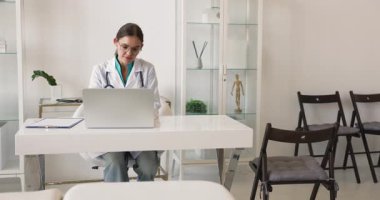 Paltolu kadın doktor iş yerinde dizüstü bilgisayarla oturuyor, orta yaşlı kadın hastaya danışmanlık yapıyor, profesyonel sağlık kontrolüne başlıyor, hastane muayenesinde belirtileri tartışıyor.