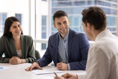 Takım elbiseli olumlu Avrupalı işadamı iş ortaklarıyla toplantı masasında oturuyor, gülümsüyor ve kalem tutuyor, sözleşme imzalıyor, müzakere sonuçlarından tatmin oluyor. Kazanç, anlaşma