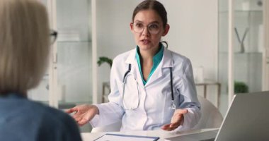 40 'lı yaşlarda beyaz önlüklü yaşlı bir kadın doktor klinikteki masada oturuyor, hastayla konuşuyor, sağlık tavsiyeleri veriyor, test sonuçlarını açıklıyor, tedavi planını paylaşıyor, tıbbi açıklamalar yapıyor.