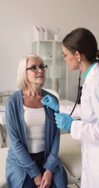 Stetoskop kullanan kadın doktor, orta yaşlı bir kadının kalp ve akciğerlerini dinliyor, hasta muayenehanesinde kalp atış hızını ve ritmi ölçüyor. Kardiyoloji, kalp hastalığı, check-up.