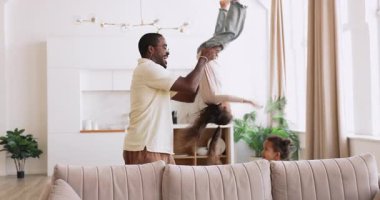 Mutlu Kara Baba, neşeli melez kızlarını baş aşağı kaldırıyor ve kanepeye oturuyor, gülüyor, modern oturma odasında birlikte oynuyor, hafta sonunu evde geçiriyor, aktif oyunların keyfini çıkarıyor. Çocuklarla ailecek vakit geçirmek