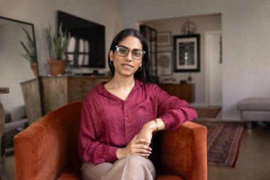 Moda gözlüklü mutlu Hintli genç ev sahibi kadın şık bir evde rahat koltukta oturuyor, kameraya bakıyor, dairede poz veriyor, keyfine bakıyor, rahatlık ve rahatlığın tadını çıkarıyor.