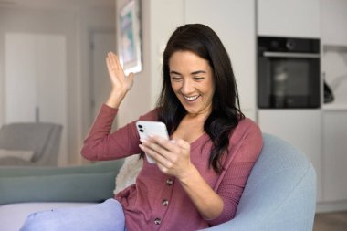 Neşeli kadın rahat bir oturma odasında oturmuş, şaşırmış bir ifadeyle akıllı telefon ekranına bakıyor, heyecanla elini kaldırırken videoda olumlu haberleri kutluyor ya da mesaj alıyor.