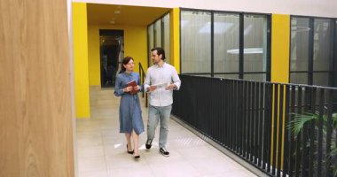 Meslektaşlar konuşuyor ve ofis koridorunda yürüyorlar, tabletteki proje detaylarını gözden geçiriyorlar, not defterindeki notları kontrol ediyorlar, işle ilgili sorunları çözüyorlar, takım çalışması, planlama, iş kararları veriyorlar.