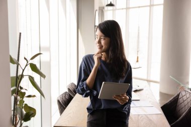 Asyalı genç ve düşünceli iş kadını elinde tabletle ofiste tek başına duruyor, çenesine dokunuyor, pencereye bakıyor, düşünüyor, internet teknolojisini iş için kullanıyor.