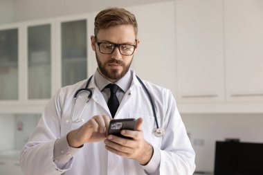 Sağlık hizmetlerinde mobil iletişim. Odaklanmış beyaz üniformalı genç erkek doktor profesyonel mesajlaşma için cep telefonu kullanıyor uzaktan konsültasyon metnini internetteki teletıp uygulaması inceleme verilerine yaz
