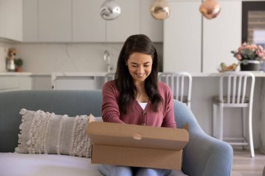 Gülümseyen güzel kadın evdeki kanepede oturuyor, karton teslimat kutusunu heyecanlı bir ifadeyle açıyor, hızlı teslimat hizmetlerinin keyfini çıkarıyor, sürpriz hediyeler alıyor. Çevrimiçi alışveriş, olumlu e-ticaret deneyimi