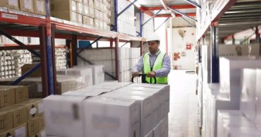 Depoda koruyucu şapkalı ve çelik yelek kamyonlu İspanyol bir erkek depo çalışanı. Endüstriyel fabrikada çalışan Latin işçi, istiflenmiş kutuları metal raflara taşıdı