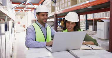 Birkaç depo çalışanı takım çalışması için laptopta buluşuyor, bilgisayar kullanıyor, çevrimiçi içeriği tartışıyor, raflardaki kutuları işaret ediyor, konuşuyor, envanter çıkarıyor.