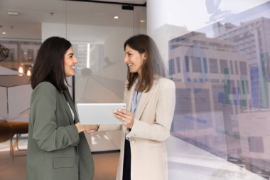 Modern ofiste duran, gülümseyen, dijital tableti tutarken el sıkışan, anlaşmayı sonuçlandıran, başarılı anlaşmayı onaylayan, üretken tartışmanın sonucunu kutlayan iki profesyonel kadın.